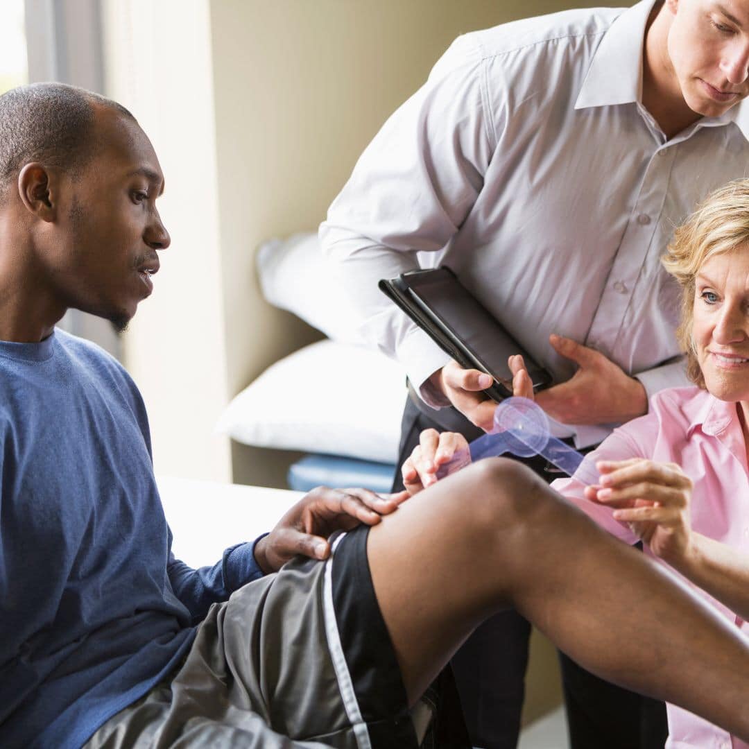 knee replacement surgery A man sits on an exam table while a healthcare professional applies a bandage to his knee and another person takes notes on a clipboard.