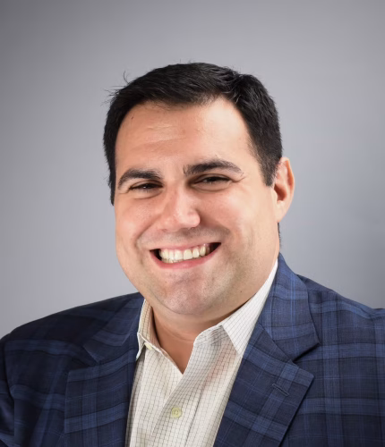 dr-mileto.jpg A man with short dark hair smiles, wearing a blue plaid suit jacket and a light-colored collared shirt, in front of a gray background.