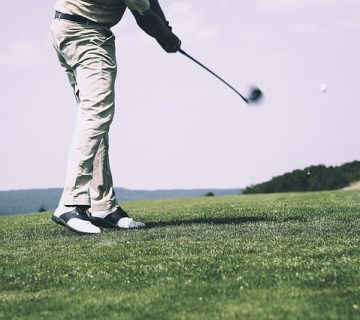 A person wearing light pants and black-and-white shoes is mid-swing with a golf club on a grassy golf course under a clear sky.