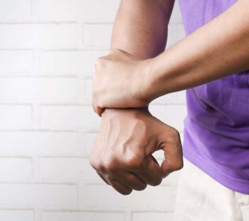 A person in a purple shirt holds their right wrist with their left hand, possibly seeking relief after a cortisone injection, standing in front of a white brick wall.