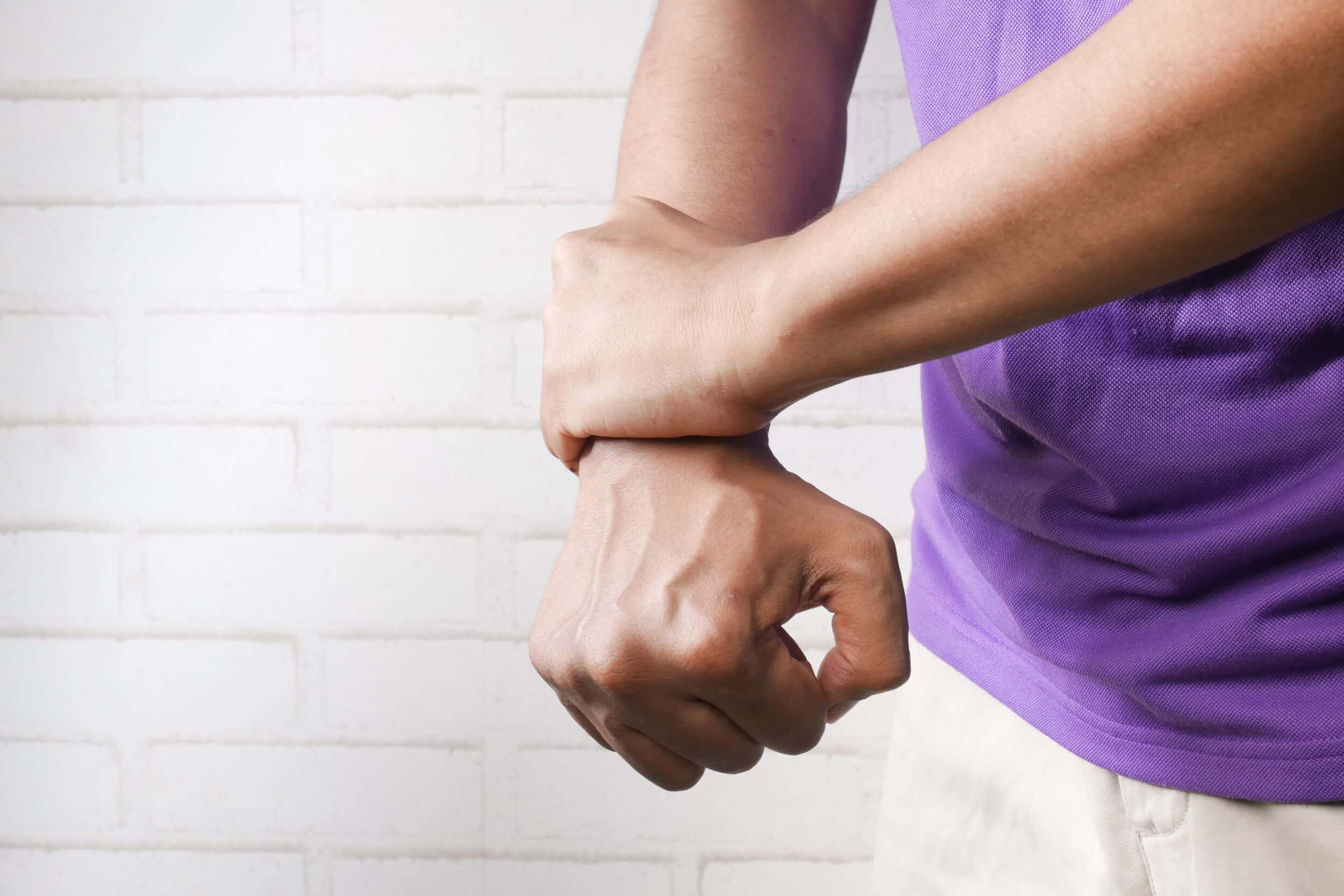 A person in a purple shirt holds their right wrist with their left hand, possibly seeking relief after a cortisone injection, standing in front of a white brick wall.