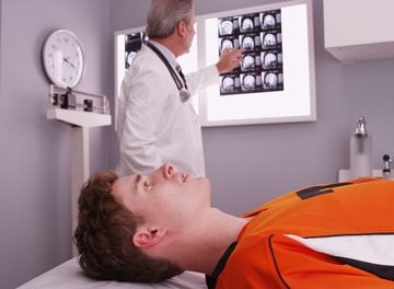A doctor examines brain scan images on a lightbox while a patient in an orange sports jersey lies on an exam table in a medical office.