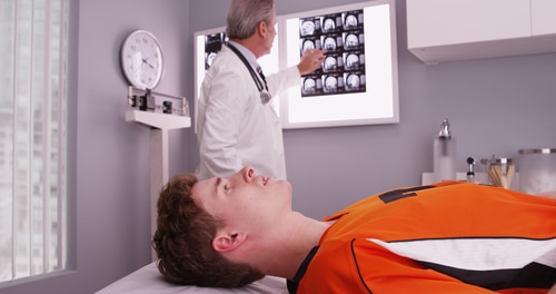 A doctor examines brain scan images on a lightbox while a patient in an orange sports jersey lies on an exam table in a medical office.