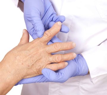 A healthcare professional wearing blue gloves examines the hand of an older adult in a red sleeve, checking for signs of arthritis.