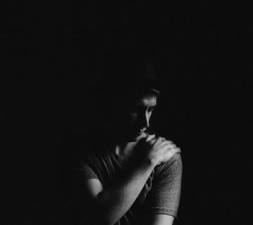 A person in a dimly lit scene looks down, with one hand resting on their opposite shoulder as if recovering from shoulder replacement surgery, all set against a dark background.
