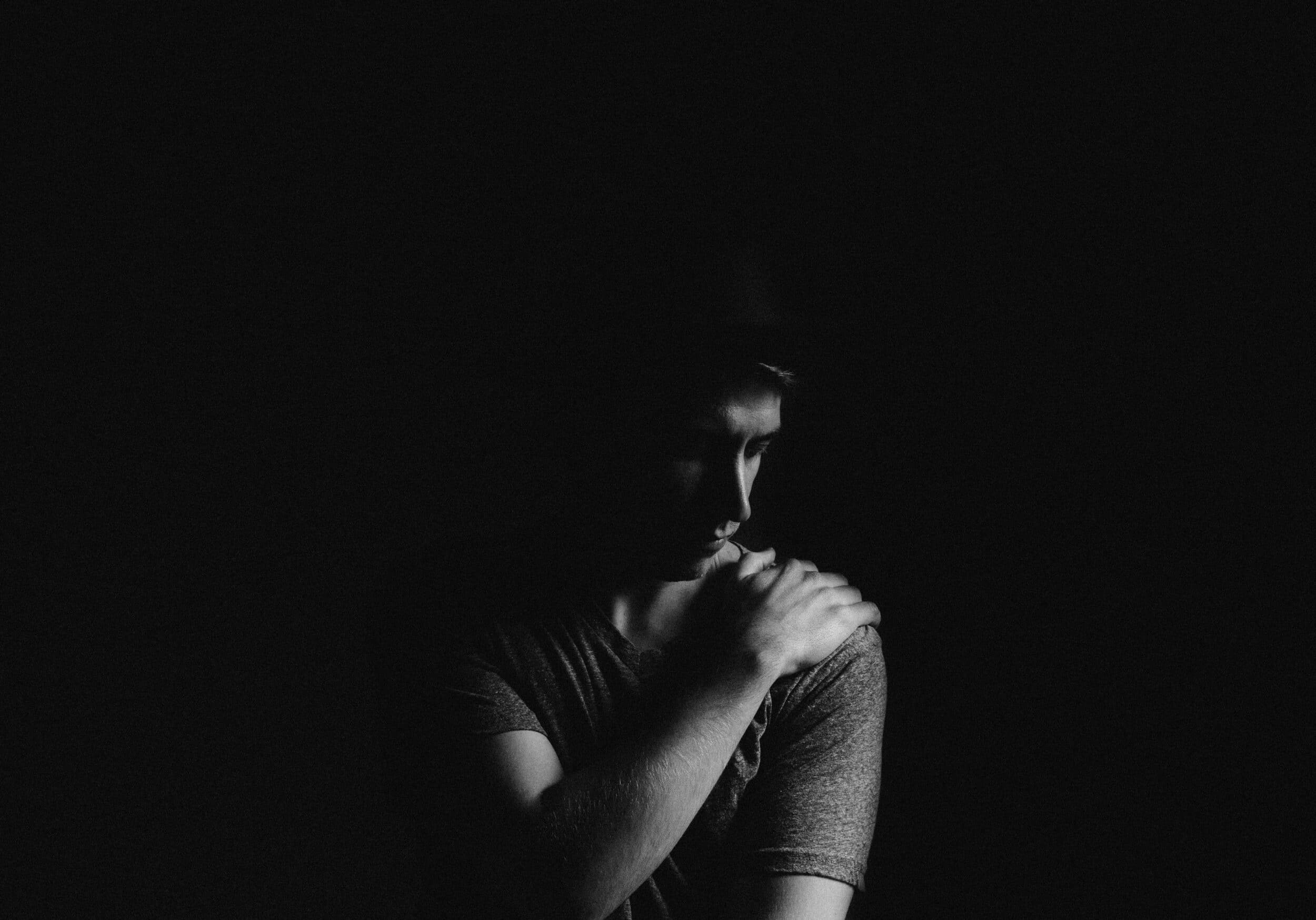 A person in a dimly lit scene looks down, with one hand resting on their opposite shoulder as if recovering from shoulder replacement surgery, all set against a dark background.