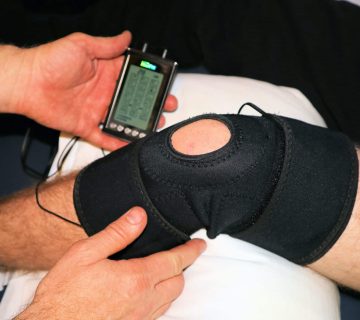 A person applies a black knee brace with electrodes connected to a portable device for physical therapy or pain management after a total knee replacement.