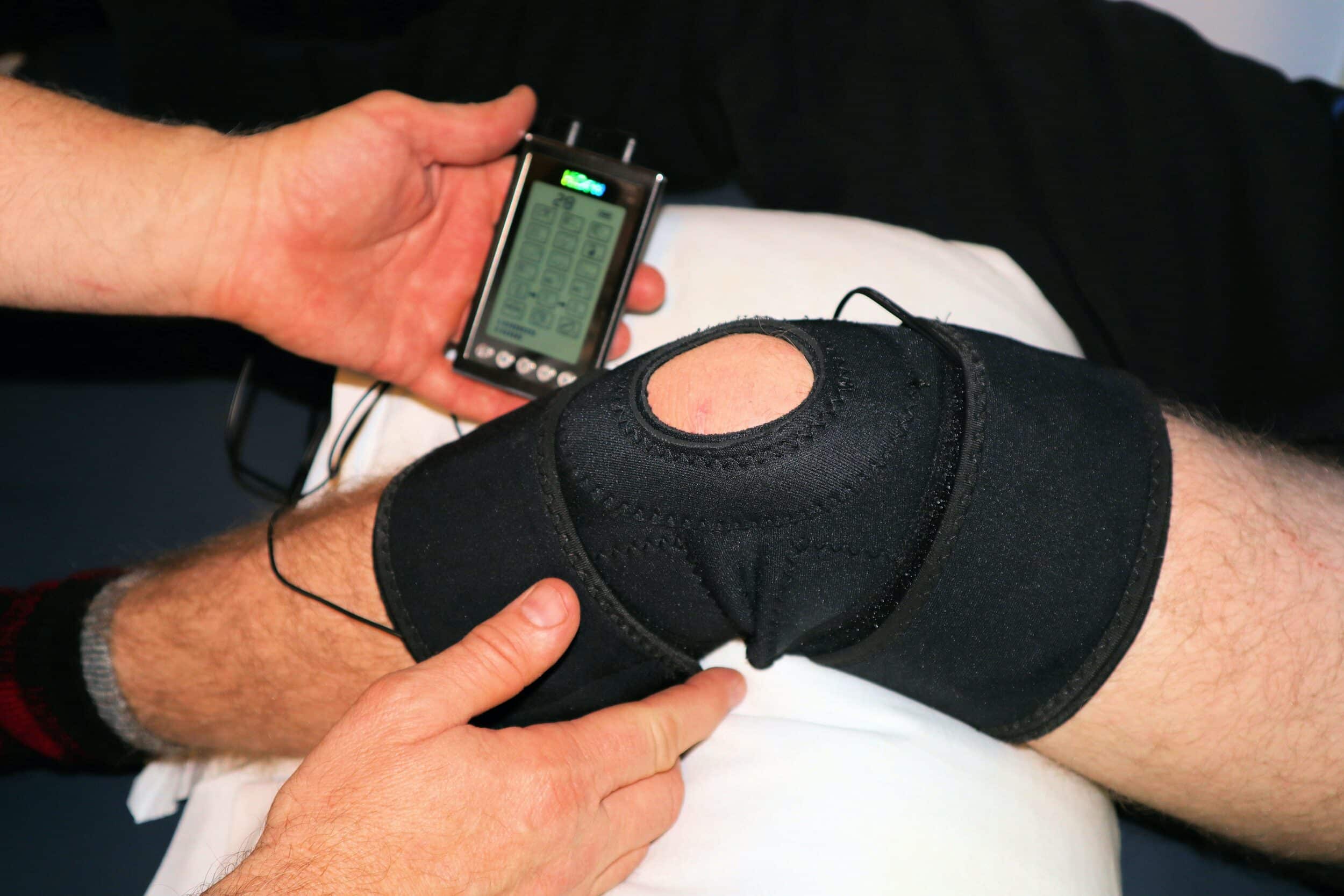 A person applies a black knee brace with electrodes connected to a portable device for physical therapy or pain management after a total knee replacement.