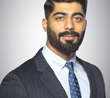 Man with a beard and neatly styled hair in a pinstripe suit and dotted tie, smiling slightly against a gray background.