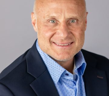 Bald man in a dark suit and blue dotted shirt, smiling at the camera against a gray background.