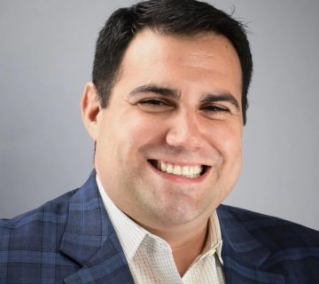 A man in a checked suit jacket and light-colored shirt smiles against a gray background.