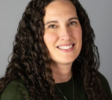 A person with long curly dark hair and wearing a dark green sweater smiles against a gray background.