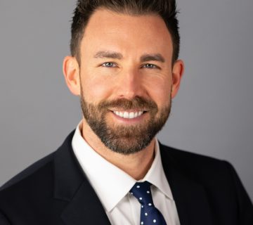 Man in a suit with a white shirt and polka dot tie smiles against a gray background.
