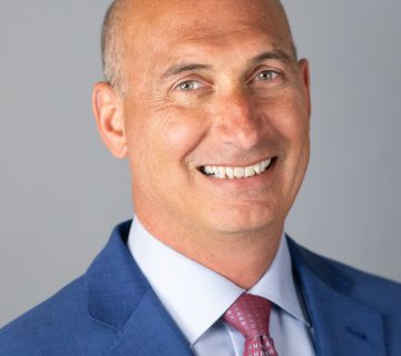 A smiling man in a blue suit and pink tie stands against a plain gray background.