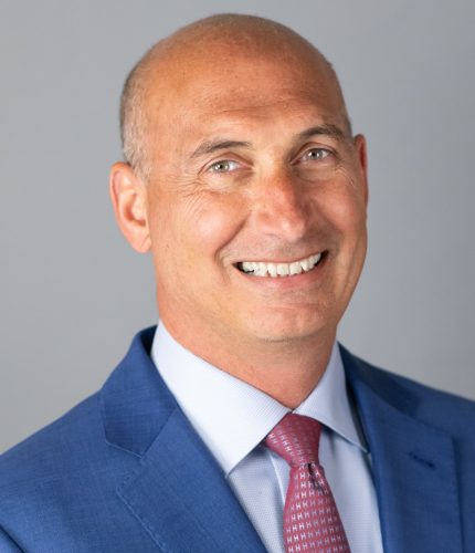 A smiling man in a blue suit and pink tie stands against a plain gray background.