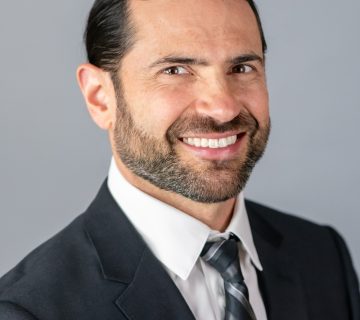 Man in a suit and tie smiles, posing against a plain gray background.