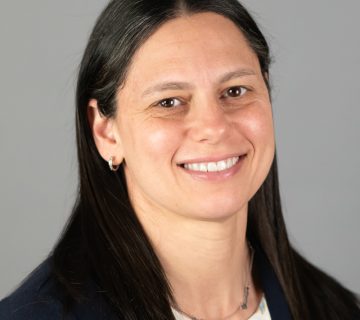 A person with long dark hair smiling, wearing a patterned blouse and dark blazer, set against a plain gray background.