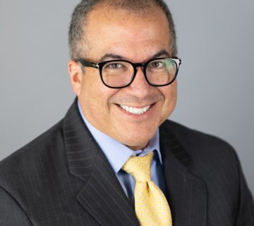 A person with glasses in a dark suit, light blue shirt, and yellow tie smiles against a plain gray background.