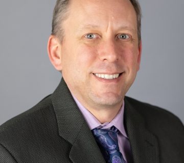 Man in a suit and tie smiles against a gray background.