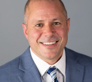 A person wearing a blue suit with a striped tie smiles against a gray background.