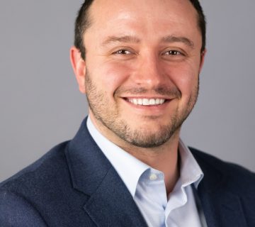 A man with short dark hair and a beard is smiling, wearing a blue suit jacket and white shirt against a plain gray background.