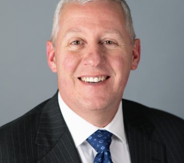 Smiling man in a dark suit with a blue patterned tie on a plain gray background.