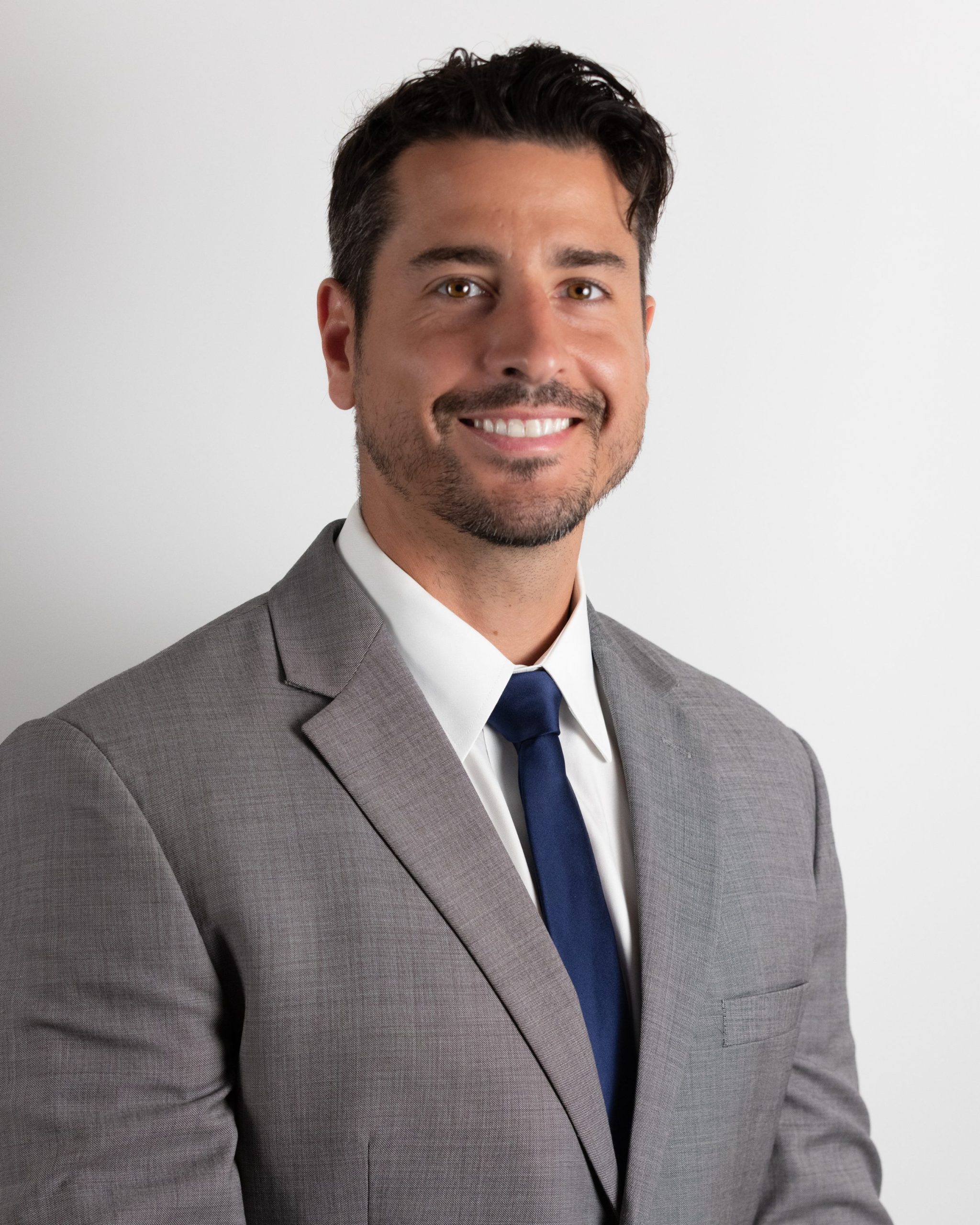 image (3) A man with dark hair and a beard, wearing a light gray suit, white shirt, and navy blue tie, smiles in front of a plain white background