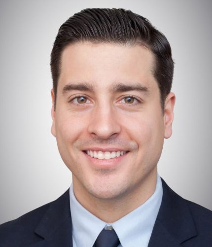 Gregory Person with short dark hair in a suit and blue tie, smiling at the camera against a neutral background.