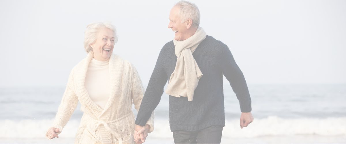 An older couple walks hand in hand on a beach, both smiling. They are dressed in warm sweaters and scarves. The ocean waves are visible in the background.
