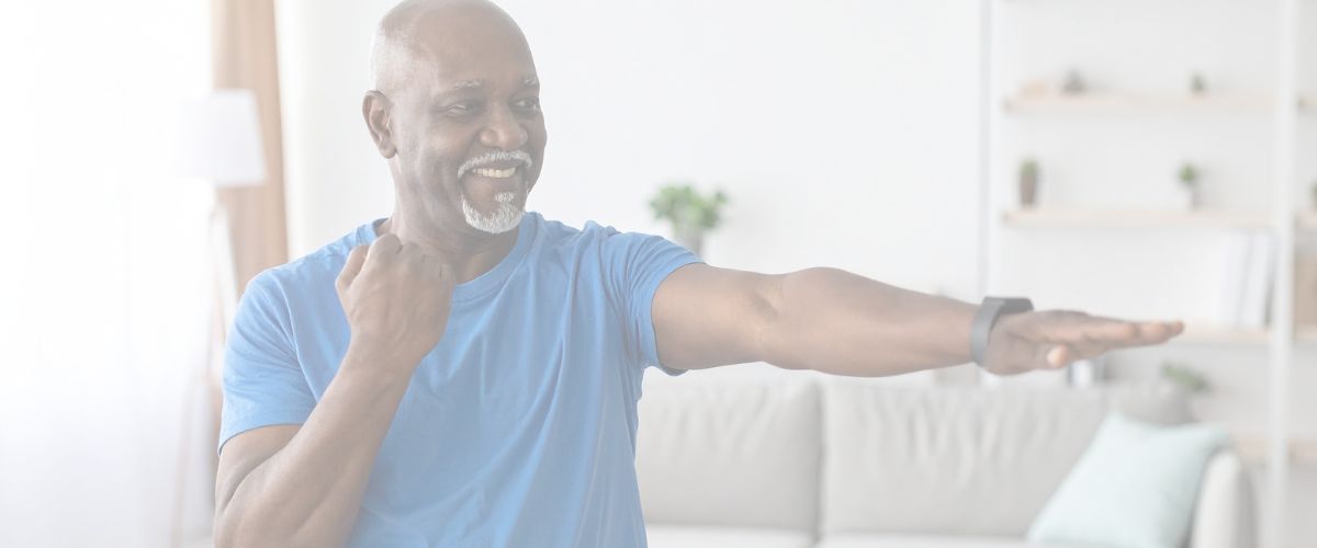 An older man in a blue t-shirt exercises indoors, smiling and extending one arm forward while holding the other arm bent near his chest.