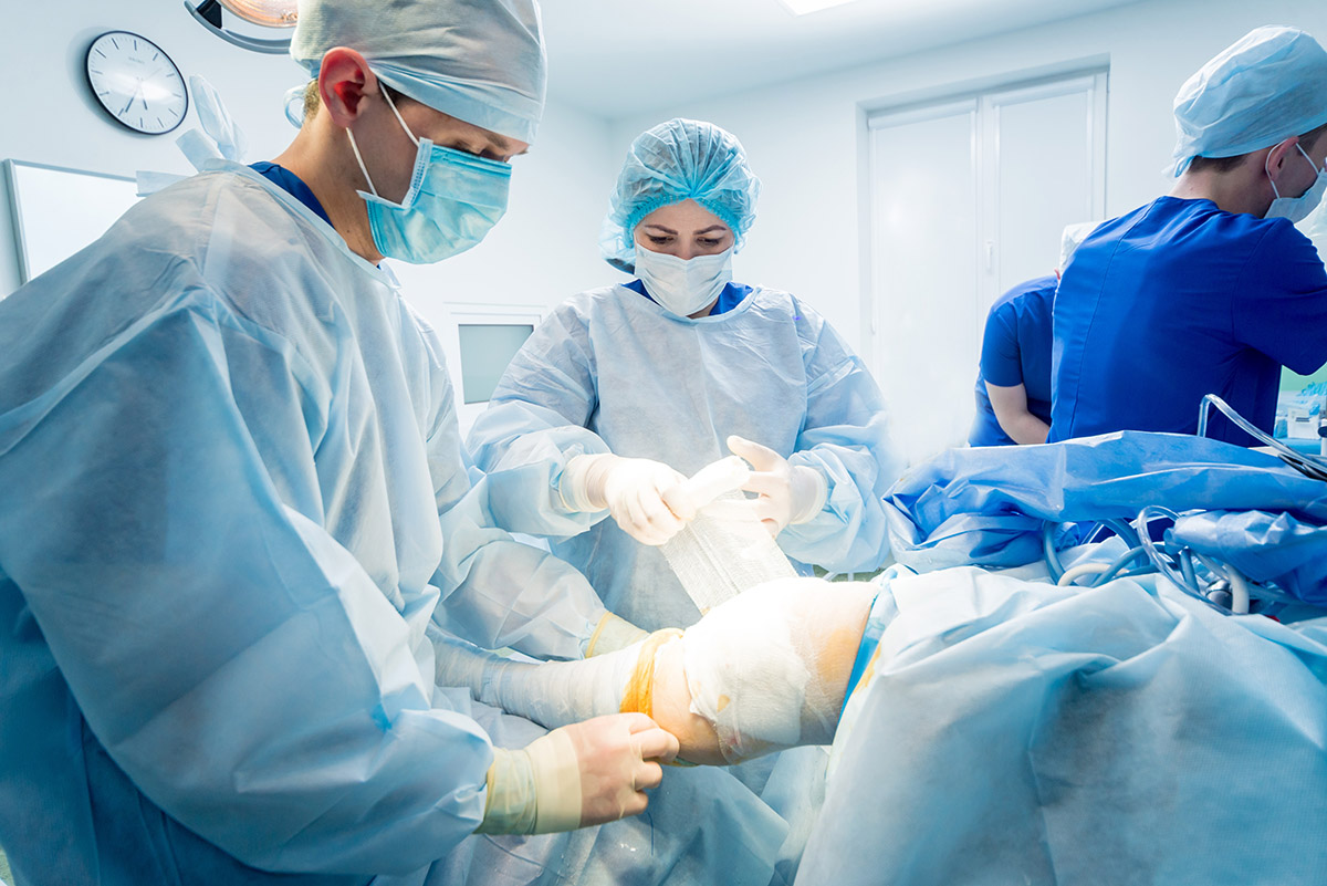 Surgeons in sterile gowns and masks perform an operation on a patient's leg in a brightly lit operating room.