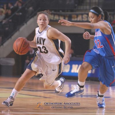 OALI LP (20) Two female basketball players are in action on the court; one in a white Navy uniform dribbles the ball while a player in a blue uniform with "12" defends
