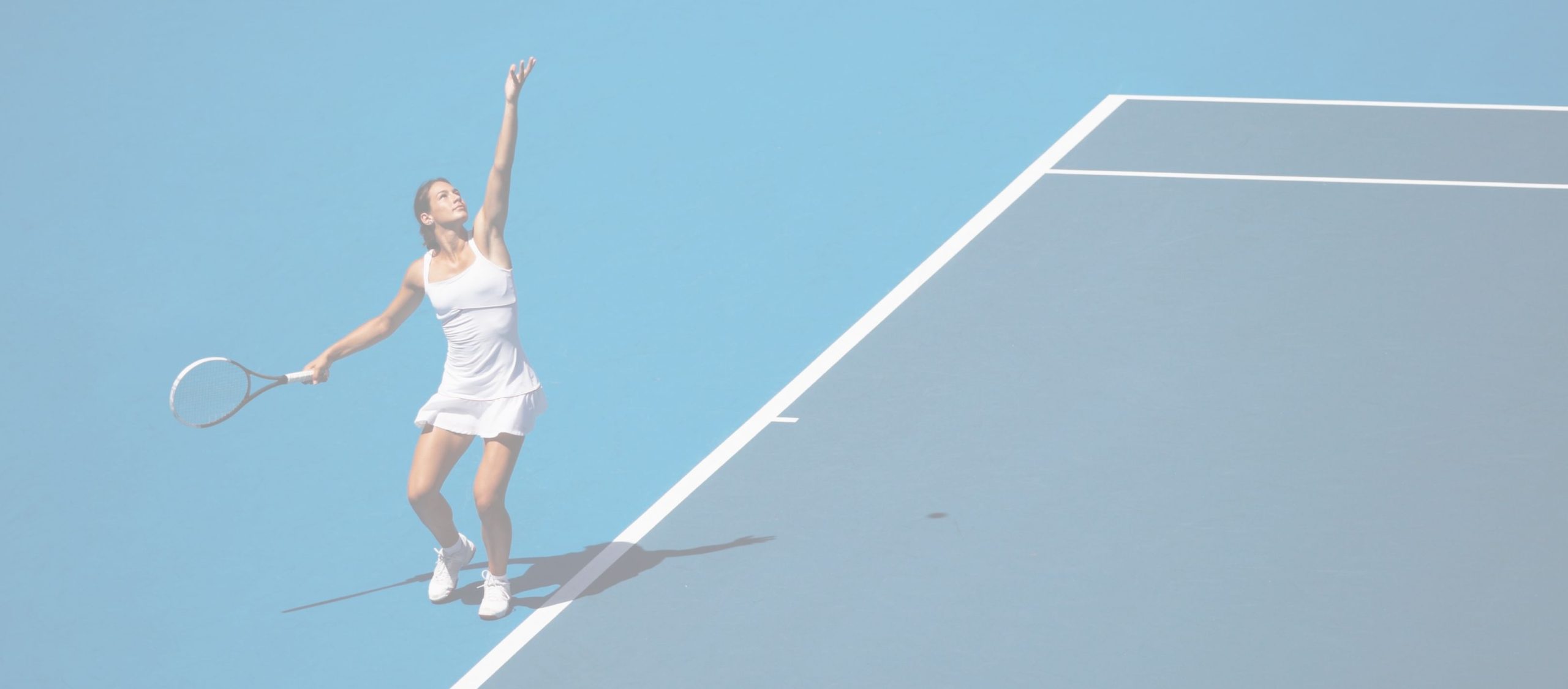 A female tennis player in a white outfit prepares to serve on a blue hardcourt, positioned near the baseline.