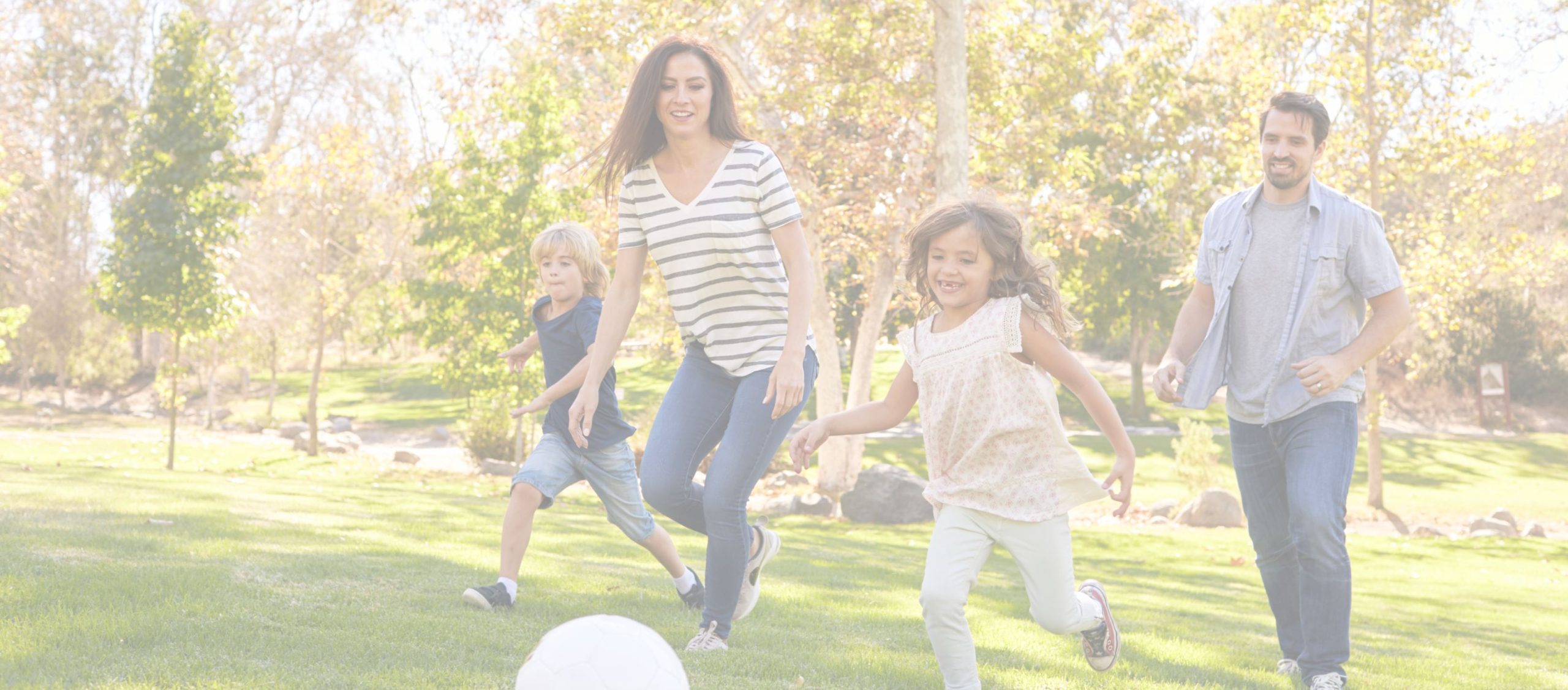 A family of four runs together on a grassy field, chasing a soccer ball on a sunny day.