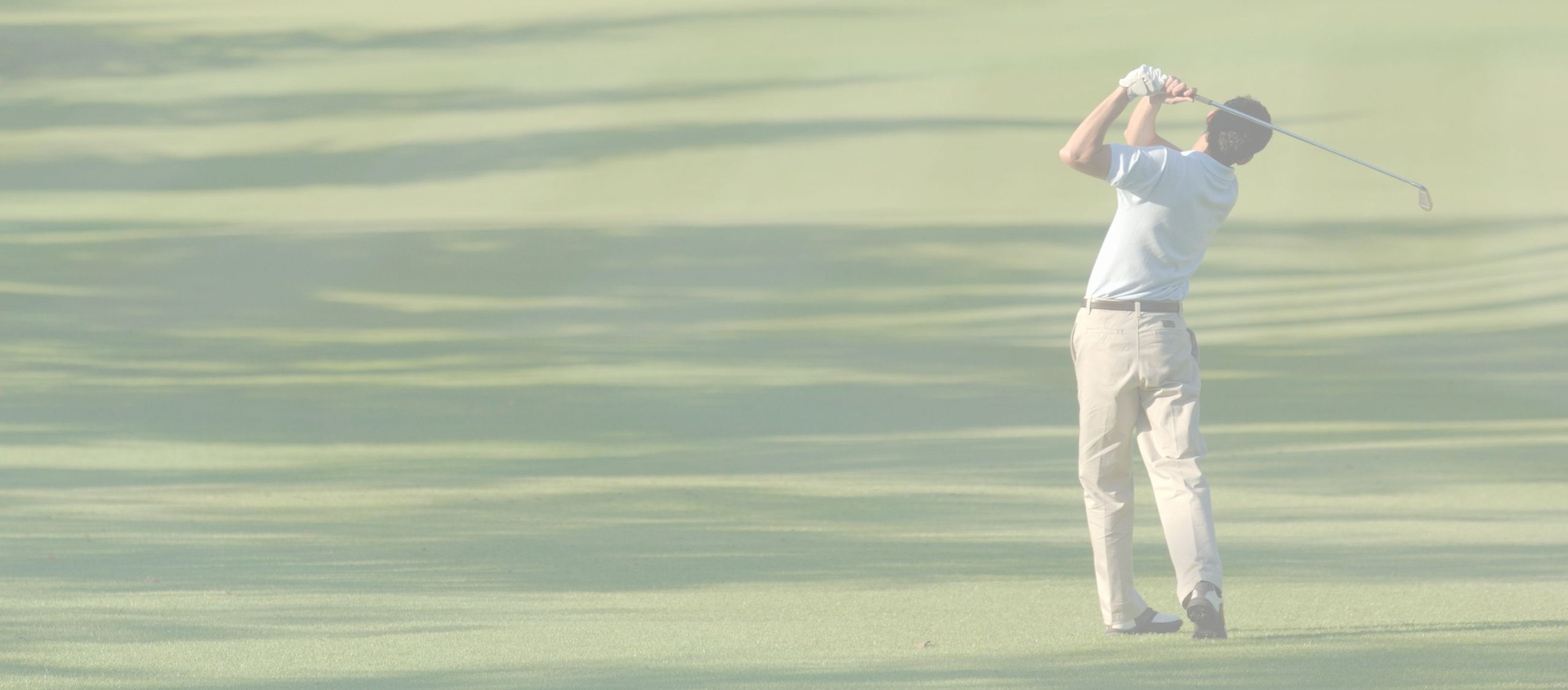 A person wearing a light shirt and khaki pants stands on a golf course, swinging a golf club toward the green.