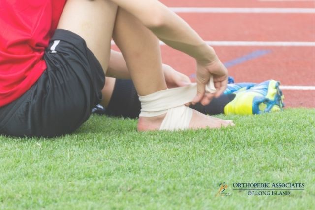 OALI LP (12) A person sitting on grass next to a running track wraps an elastic bandage around their ankle