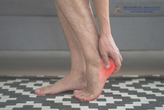 OALI LP (13) Person holding their heel, with a red area indicating pain or inflammation, standing on a patterned rug "Orthopedic Associates of Long Island" logo appears in the top right corner