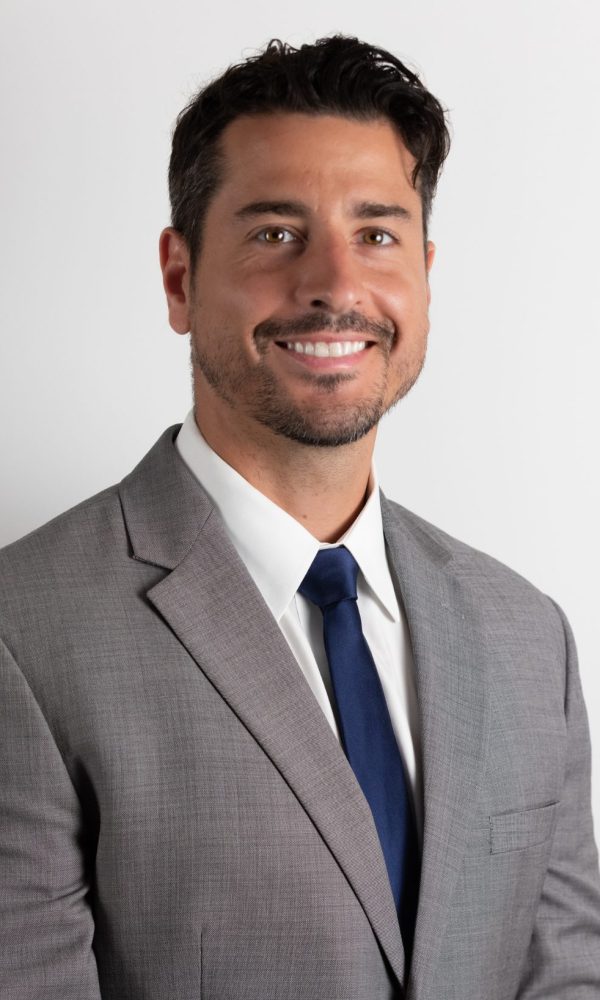 Untitled design (17) Man in a light gray suit, white shirt, and dark blue tie smiles in front of a plain white background
