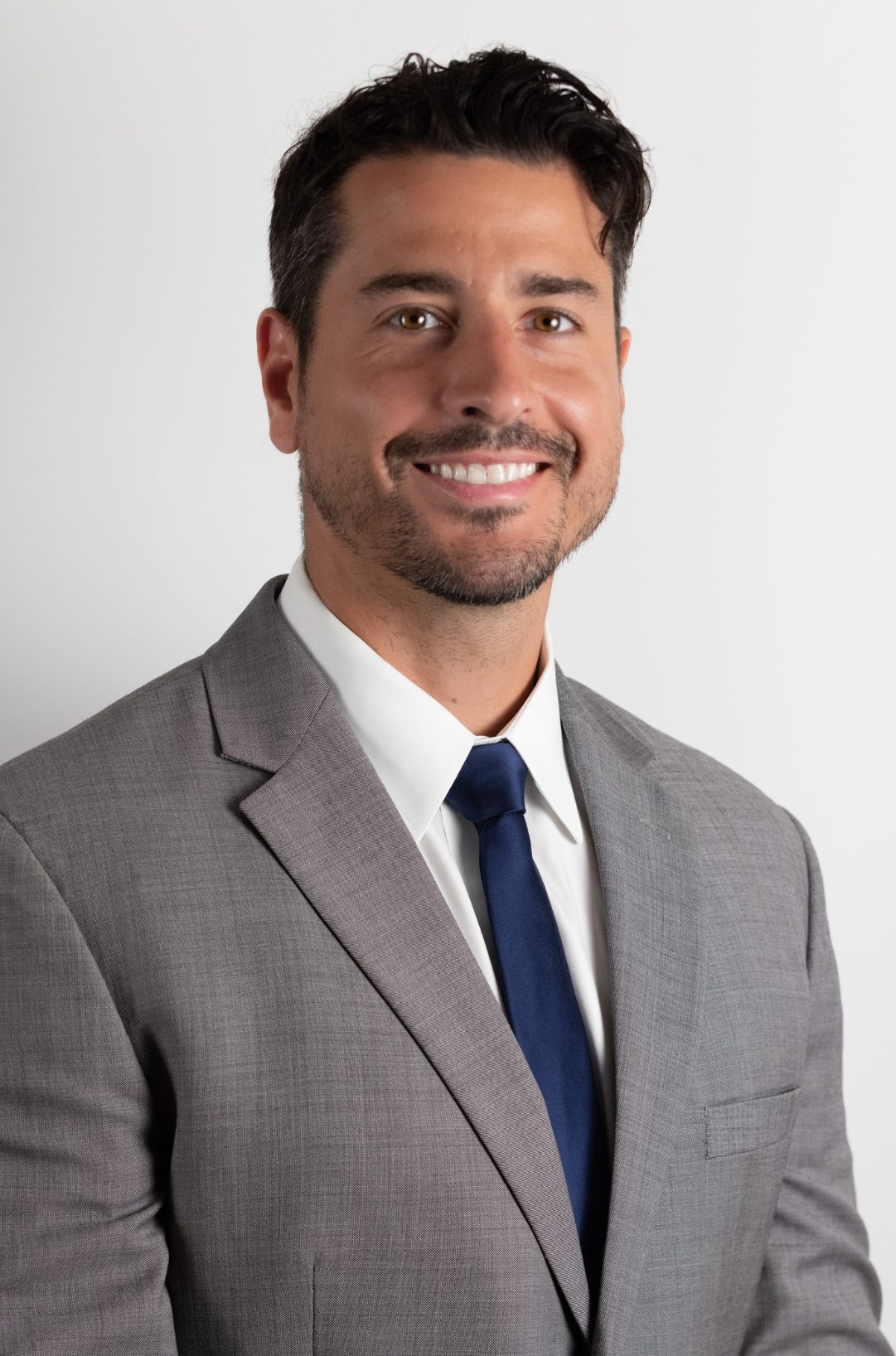 Man in a light gray suit, white shirt, and dark blue tie smiles in front of a plain white background 